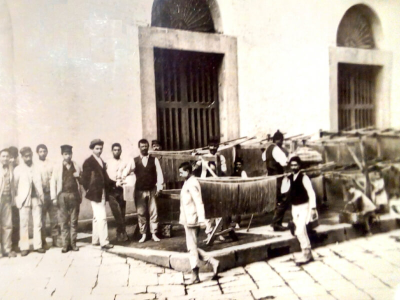 An old sepia coloured image of a group of men in Italy on a stone street in front of a building with arched windows